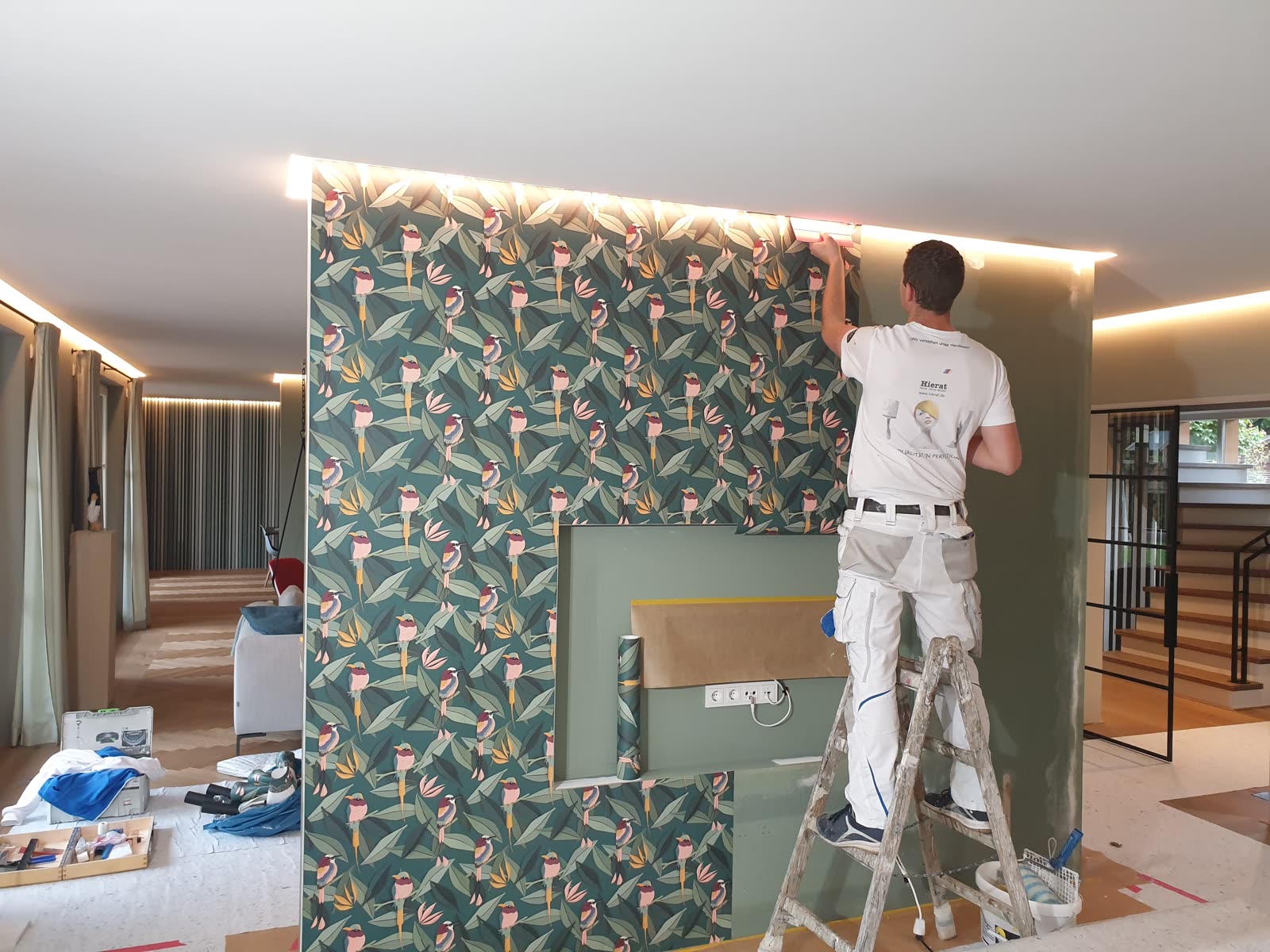 Hierat painter decorating a living room wall with green bird wallpaper